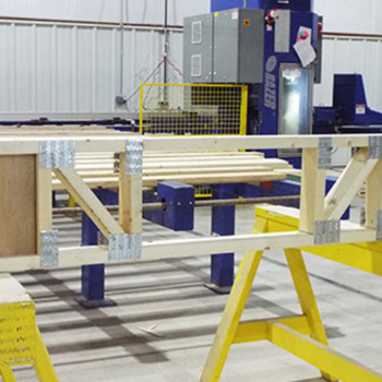 A woodworking setup with wooden planks on a table supported by yellow sawhorses, and a machine in the background.