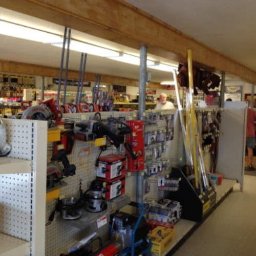 Hardware store interior with shelves displaying tools like saws, paint supplies, and other equipment. A person is browsing in the background.