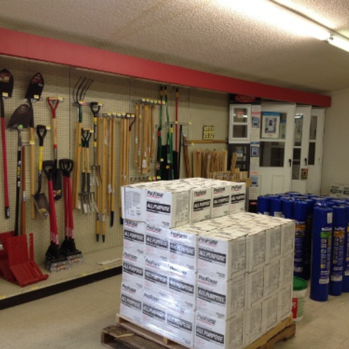 A hardware store interior with various tools like shovels and rakes on a wall, a pallet of boxed products on the floor, and blue rolls nearby.