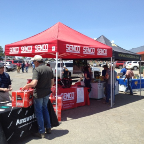Outdoor trade show with a red Senco tent displaying tools, attended by people browsing and engaging. Sunny day, vibrant and busy atmosphere.
