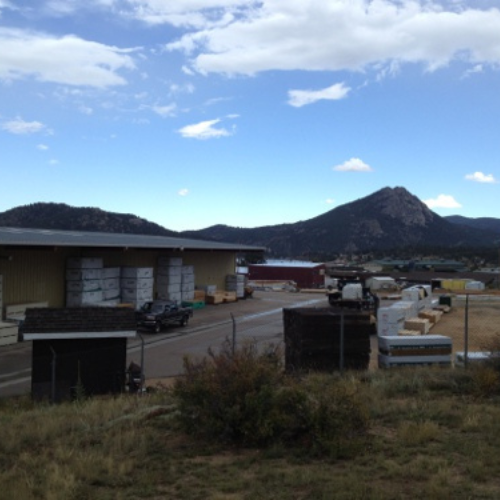 Industrial yard with stacked materials and a warehouse, set against a mountainous backdrop under a bright, partly cloudy sky. Quiet and expansive.