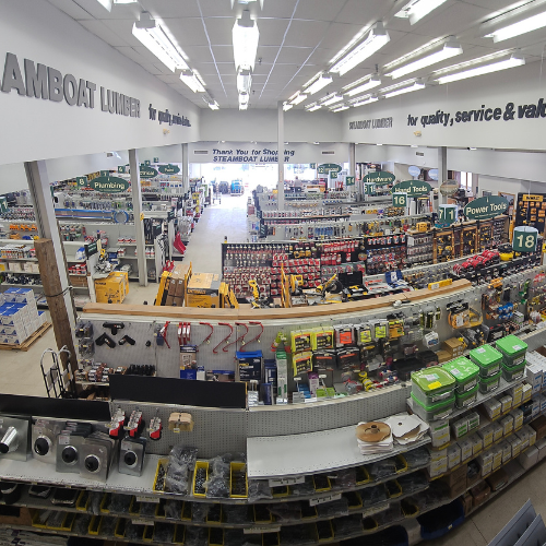 A bright, organized hardware store with aisles full of tools, paints, and supplies. Overhead signs guide customers to sections like plumbing and power tools.