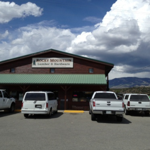 A rustic building with a green roof labeled 'Rocky Mountain Lumber & Hardware,' set against a backdrop of mountains and a cloudy sky.