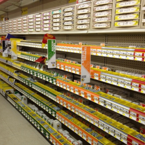 A hardware store aisle with neatly arranged shelves of labeled boxes containing various screws and fasteners, organized by size and type, under fluorescent lights.