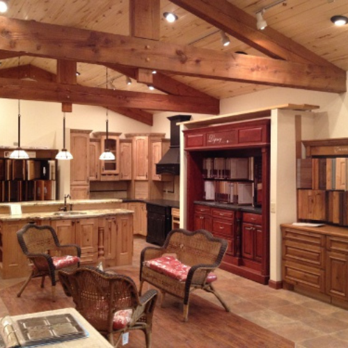 Rustic kitchen showroom with wooden cabinets and stone countertops. Warm tones, wicker chairs, pendant lights, and exposed wooden beams create a cozy atmosphere.