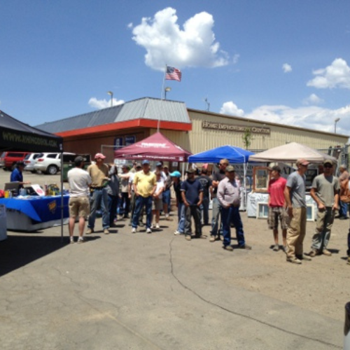 People stand in line under blue skies at an outdoor event near a home improvement center. Tents provide shade, and an American flag is visible.