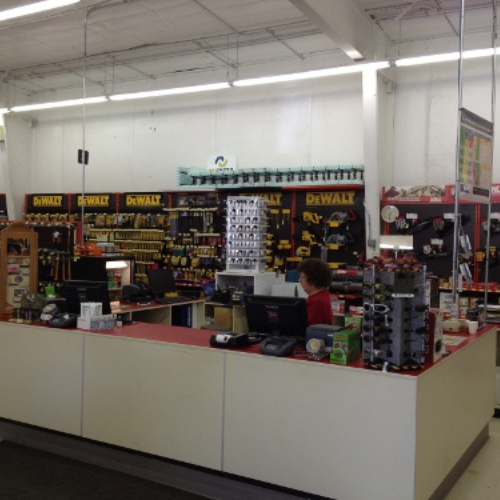Hardware store interior with a counter, showcasing various DeWalt tools on shelves. A person stands behind the register, and displays of accessories are visible.