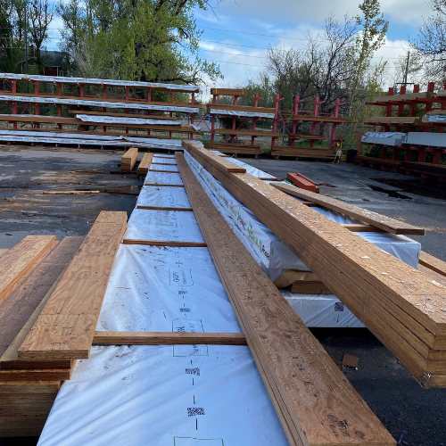 Stacks of long wooden planks and boards are neatly arranged on lined timber racks outdoors on a clear day, surrounded by green trees and cloudy skies.