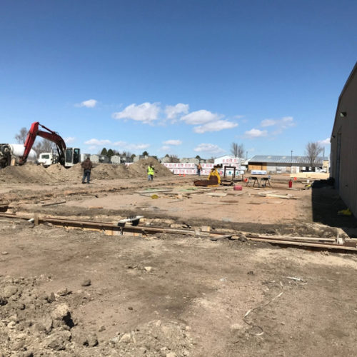 Construction site with dirt piles and machinery under a clear blue sky. Workers wearing safety gear are scattered around the area.