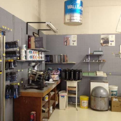 A small paint supply shop with shelves holding paint cans, brushes, and tools. A paint mixer and safety posters contribute to an organized, industrial feel.