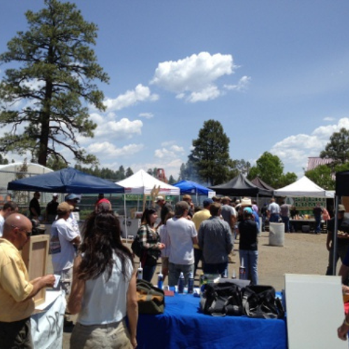 Outdoor market scene with people browsing under blue and white tents on a sunny day. Tall trees and fluffy clouds in the background create a relaxed atmosphere.