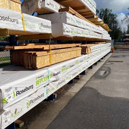 Stacks of wooden beams wrapped in Roseburg branding are neatly arranged on blue metal shelves at an outdoor construction supply yard.