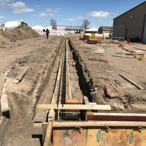 Construction site with a deep trench lined with wooden supports, surrounded by equipment and materials. A person walks in the background under a blue sky.