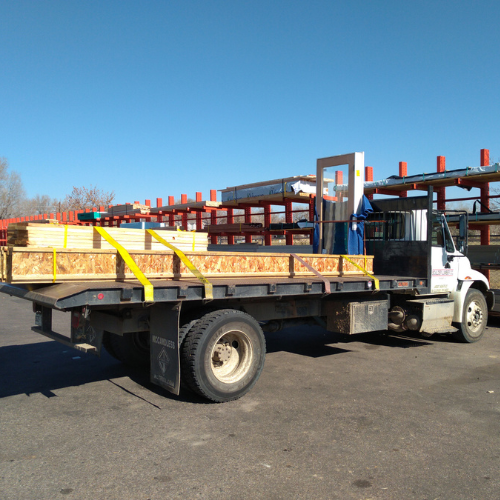 Flatbed truck parked in a lumber yard, loaded with wooden panels secured by yellow straps. Clear blue sky and red metal racks in the background.