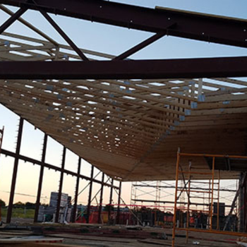 A partially constructed building showing a wooden roof framework and metal support beams, with scaffolding in the foreground and a sunset in the background.