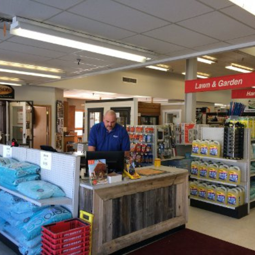 A hardware store's interior with a man in a blue shirt working at a wooden counter. Nearby are shelves with lawn and garden products, including soil bags and fertilizers. The setting is organized and functional, conveying a sense of productivity.