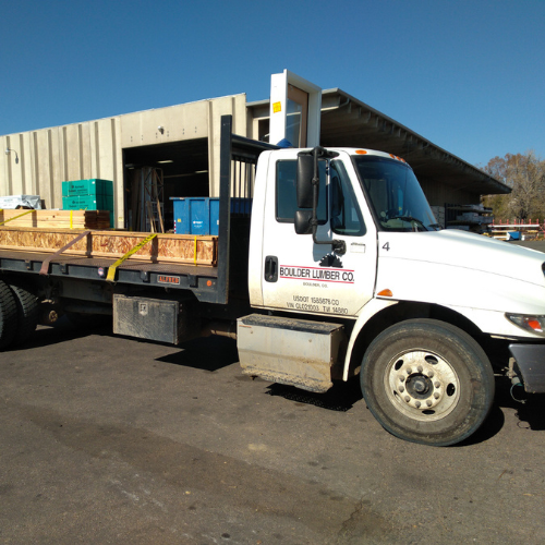 A white flatbed truck from Boulder Lumber Co. is parked beside a warehouse. The truck's bed is loaded with wooden crates. The scene is sunny.