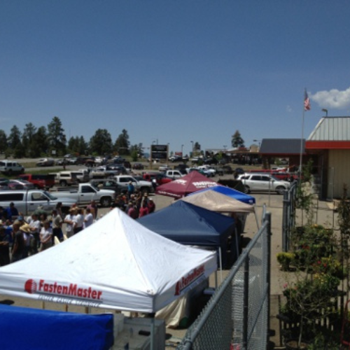 Outdoor market scene with colorful vendor tents and people browsing under a clear blue sky. Cars are parked nearby, and an American flag is visible.