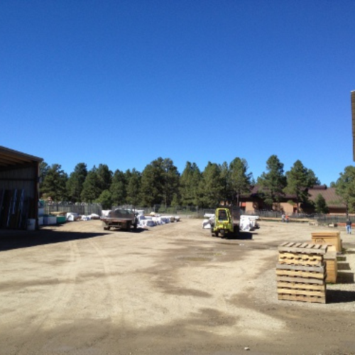 A construction yard with stacked pallets and vehicles under a clear blue sky. Surrounded by trees, the atmosphere is industrious and calm.