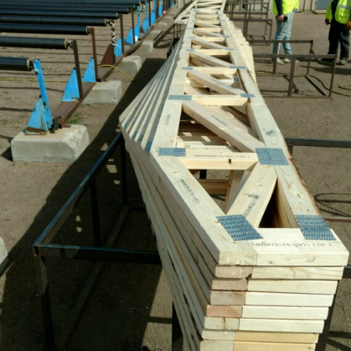 Stack of wooden trusses neatly arranged on metal stands at a construction site. Workers in safety vests are visible in the background, suggesting an active work environment.