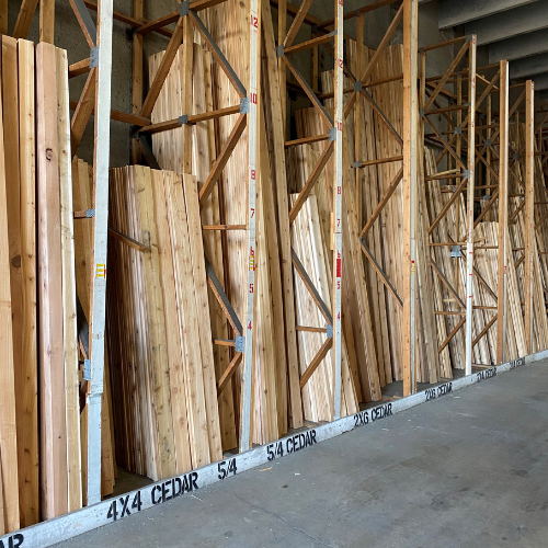 Stacks of cedar wood planks neatly organized in a warehouse. Labels on the floor indicate sizes like 4x4 and 5/4. The scene suggests a well-maintained lumber supply.
