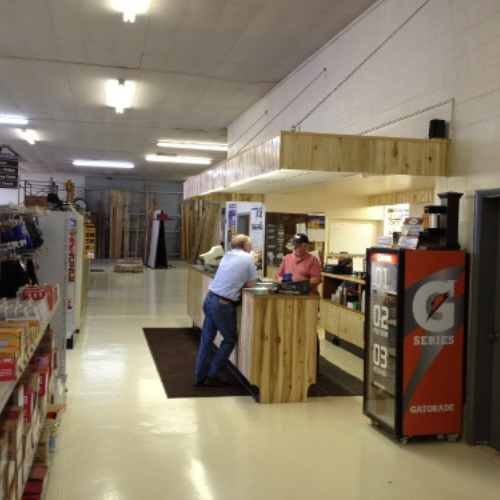 A customer talks to a cashier at a wooden counter in a bright hardware store. Shelves with products and a Gatorade vending machine are visible.