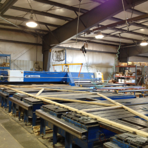 Industrial warehouse interior showing a large blue CNC machine on the left, metal beams, stacks of wooden planks, and shelving with tools, conveying a busy work environment.