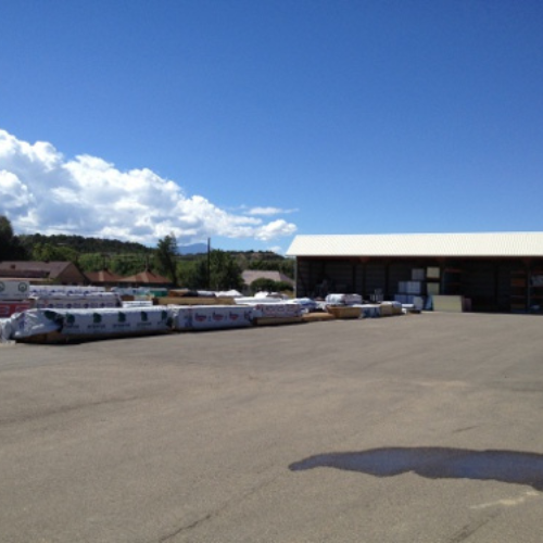 A paved outdoor storage area with stacks of packaged goods, a large warehouse, and a clear blue sky with fluffy clouds, conveying a calm, sunny day.