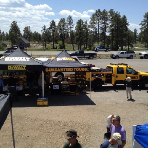 Outdoor scene featuring a DeWalt promotional setup with branded tents and a yellow truck. Trees and blue sky in the background; people browsing.