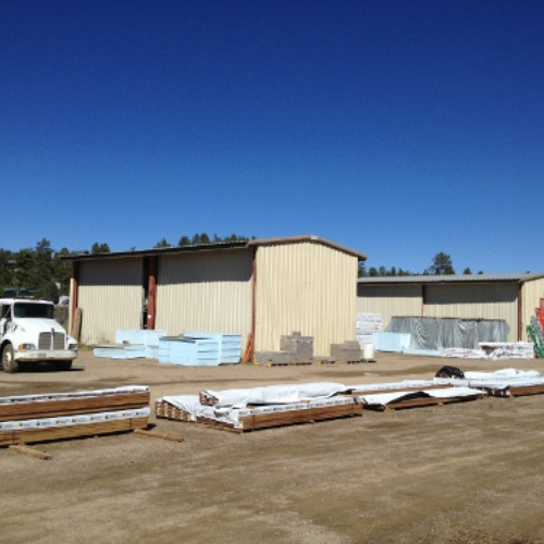 Industrial yard under a clear blue sky, featuring beige warehouses, a parked white truck, and stacks of construction materials on the ground.