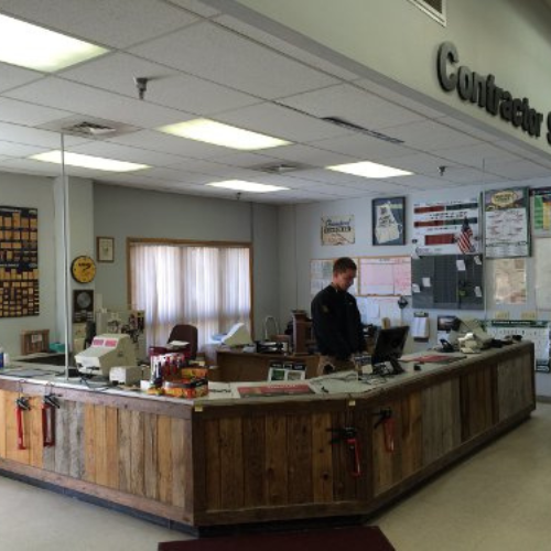 A man stands behind a rustic wooden counter in a contractor's office, surrounded by paperwork and office supplies. The atmosphere is busy and organized.