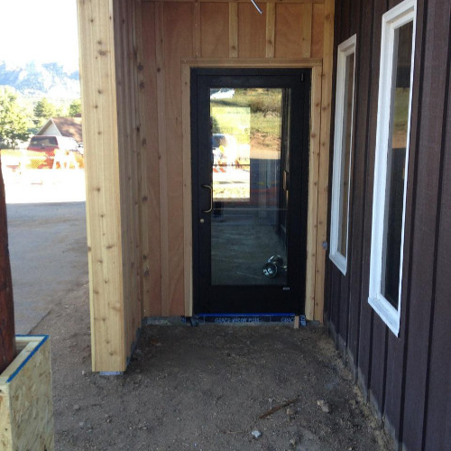 Glass door opening directly onto a dirt path, flanked by tan and brown wooden walls. A reflection of trees and sky is visible on the door.