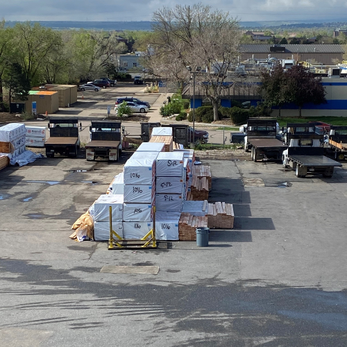 A lumber yard with stacked wooden planks covered in white tarps marked with numbers. Trucks are lined up in the background, with a few trees visible.