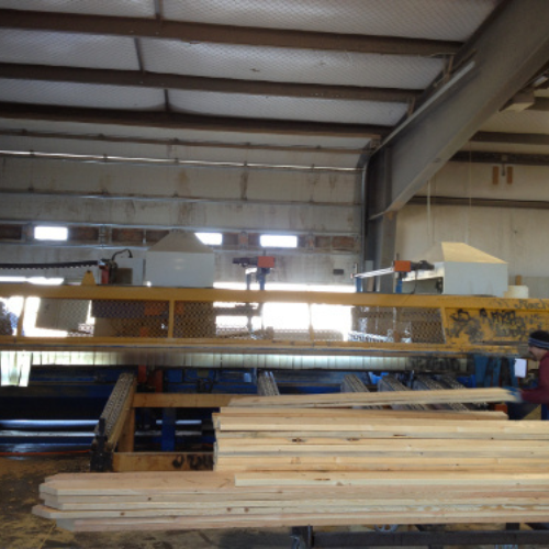 Interior of a lumber mill with stacks of wooden planks in the foreground. Industrial machinery and beams create a busy, functional atmosphere.