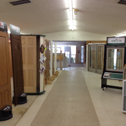 A hallway in a showroom displays various wooden doors and glass window panels. The setting is well-lit, clean, and organized, conveying a professional tone.