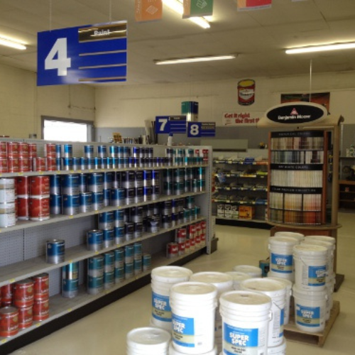 A store interior with aisles of paint cans; shelves display red and blue paint cans on the left, while white buckets are stacked on the floor. Signs with numbers 4, 7, and 8 hang overhead.