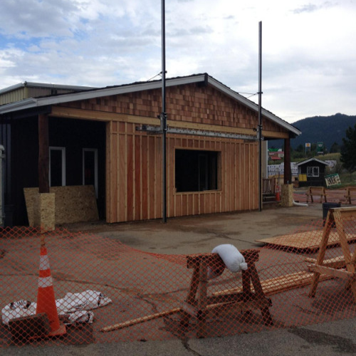 Construction scene with a partly built wooden structure, orange safety netting, and traffic cones. A person is sawing wood in sunlight.