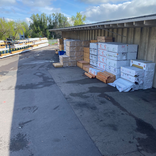 Outdoor storage area with stacked boxes and wooden pallets under an overhang on a sunny day. Trees and blue sky are visible in the background.