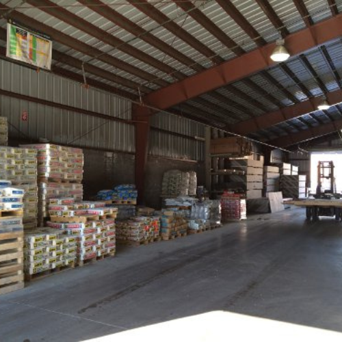 A spacious warehouse interior with stacks of boxed goods on pallets along the wall, lit by overhead lights, creating an organized, industrious atmosphere.