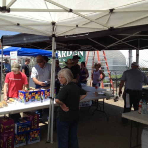 Outdoor event under tents with several people, food tables, and snacks. Attendees chat and serve themselves under sunny skies, creating a lively, friendly atmosphere.