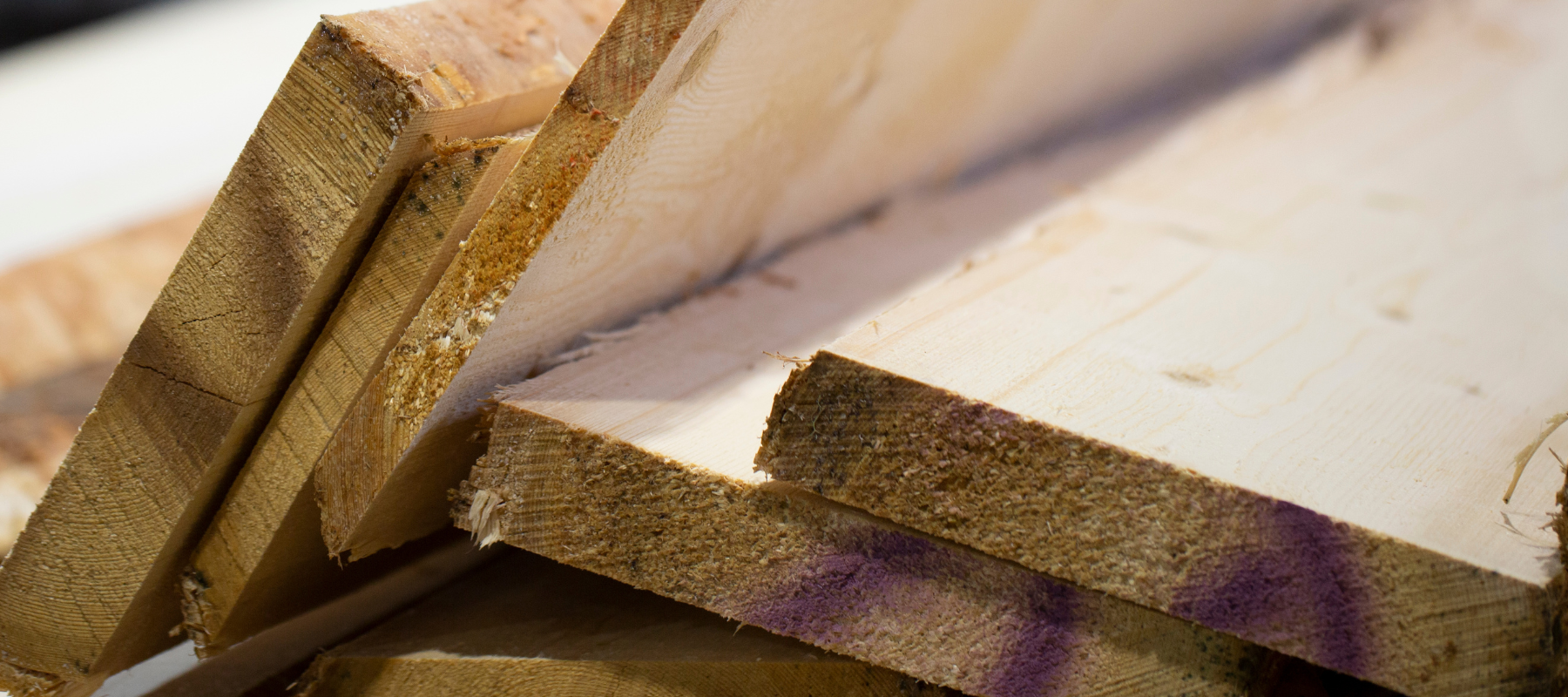 Stack of unfinished wooden planks with rough edges, leaning at an angle. The wood shows natural grain patterns, with purple marks on the surfaces.