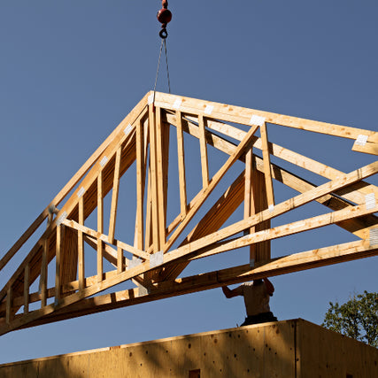 FAQsA wooden roof truss is being hoisted by a crane against a clear blue sky, while a worker stands below, guiding it onto a building. Construction scene.
