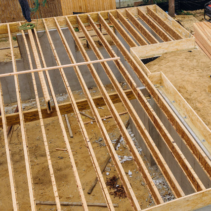 Floor Trusses & SCS TrueTrimWooden beams are arranged in a grid pattern over a sandy construction site, suggesting an early stage of building a structure. The scene conveys progress.